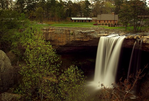A serene waterfall cascades over a rocky ledge, surrounded by lush greenery and a rustic building in the background.