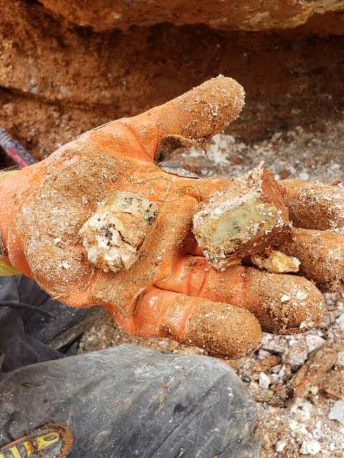 A gloved hand holds two small, rough stones covered in dirt, with a rocky background.