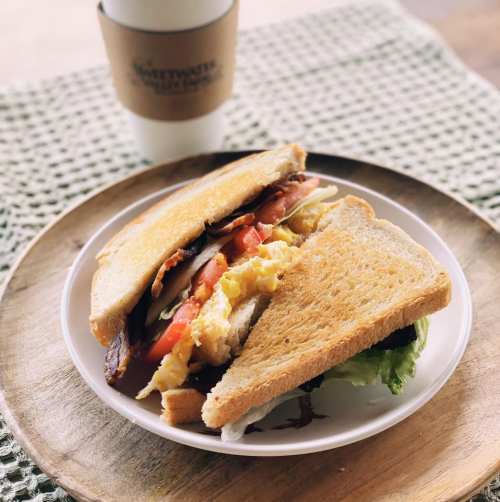 A toasted sandwich filled with bacon, eggs, tomatoes, and lettuce, served on a wooden plate with a coffee cup in the background.