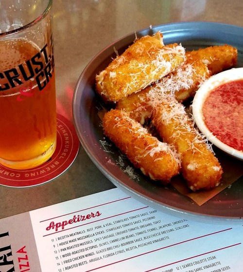 A plate of crispy fried cheese sticks with marinara sauce, a pint of beer, and a menu on the table.