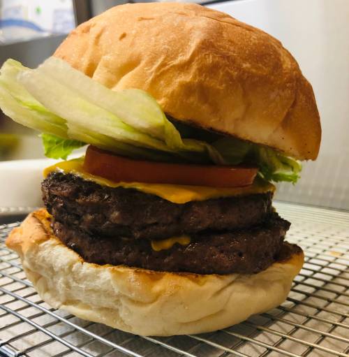 A stacked burger with two beef patties, cheddar cheese, lettuce, and tomato on a toasted bun, resting on a cooling rack.