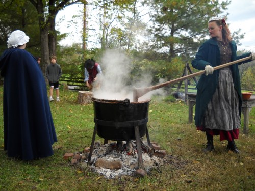 A woman stirs a large pot over a fire, surrounded by people in historical clothing in a wooded outdoor setting.