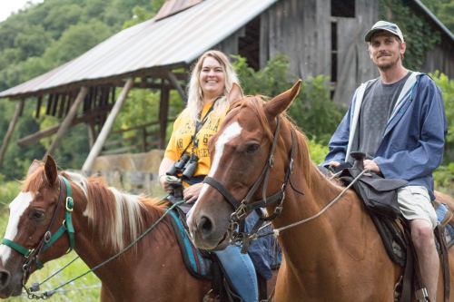 Two people riding horses near a rustic barn, smiling and enjoying a leisurely outdoor experience.