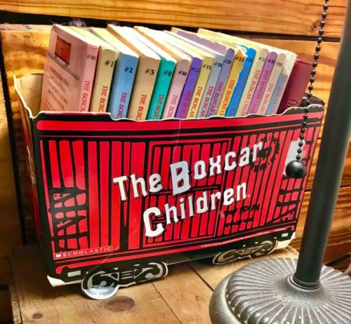 A colorful boxcar-shaped container holds a collection of "The Boxcar Children" books on a wooden shelf.
