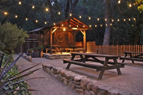 A rustic outdoor gathering space with picnic tables, string lights, and a wooden shelter surrounded by trees at dusk.
