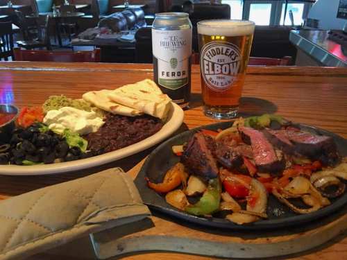 A sizzling platter of fajitas with steak, peppers, and onions, served with sides of black beans, guacamole, and drinks.