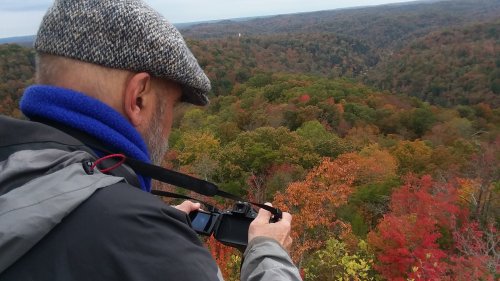 A person in a gray cap takes a photo of a colorful autumn landscape with trees in shades of red and orange.