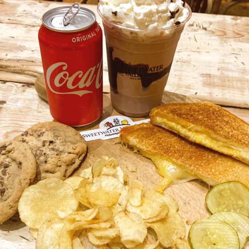 A wooden platter with grilled cheese, chips, pickles, cookies, a Coca-Cola can, and a chocolate milkshake with whipped cream.