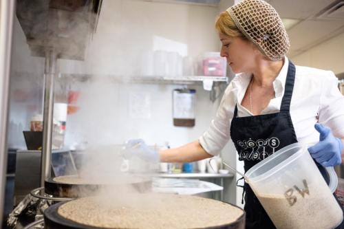 A woman in a kitchen wearing a hairnet and gloves stirs a large pot, steam rising around her.