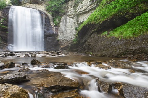 A serene waterfall cascades over rocky cliffs into a tranquil stream, surrounded by lush greenery.