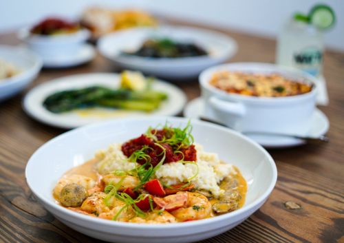 A bowl of shrimp and grits topped with sauce and green onions, with various dishes in the background on a wooden table.