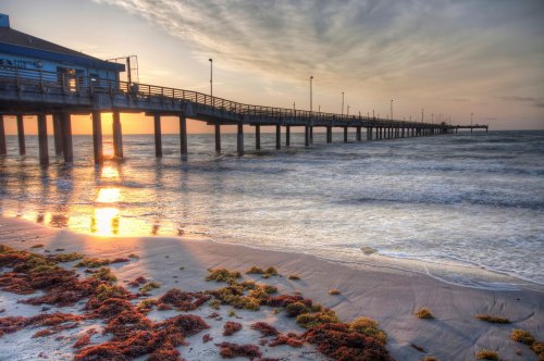 A pier extends over the ocean at sunset, with gentle waves and seaweed on the sandy shore.