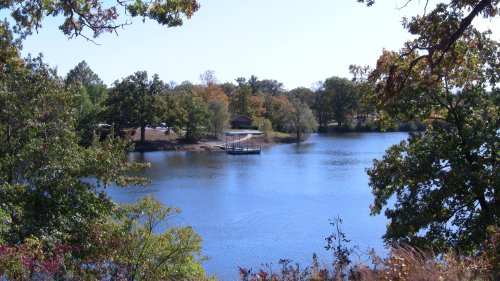 A serene lake surrounded by trees, with a small dock and a house visible on the shore under a clear blue sky.