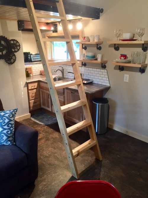 A cozy kitchen with wooden shelves, a ladder leading to a loft, and a sofa in a small, well-lit space.