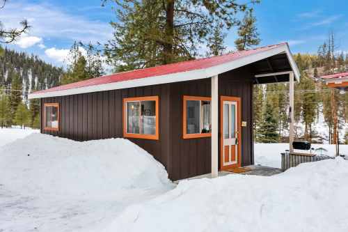 A small brown cabin with orange trim and a red roof, surrounded by snow and pine trees under a blue sky.