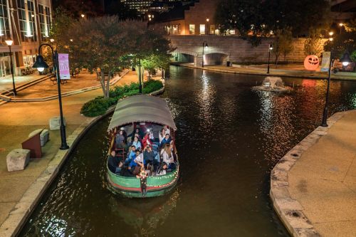 A boat with passengers glides along a lit canal at night, surrounded by trees and buildings, with a Halloween decoration nearby.