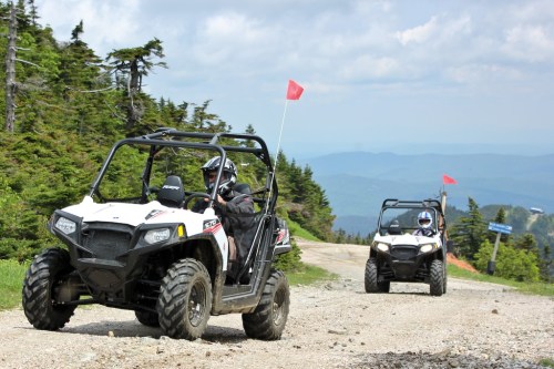 Two off-road vehicles drive along a gravel path in a forested area, with mountains visible in the background.