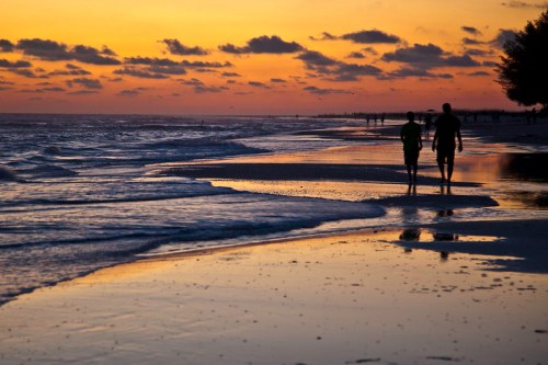 Silhouetted figures walk along a beach at sunset, with waves gently lapping at the shore and colorful clouds above.
