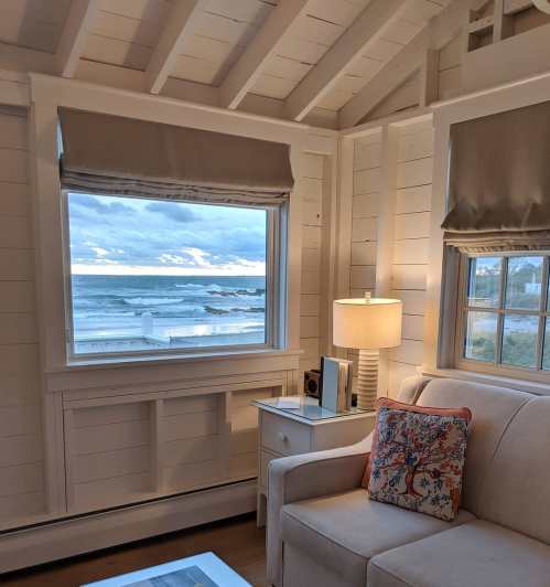 Cozy interior with a sofa, lamp, and a large window showcasing a view of the ocean and cloudy sky.