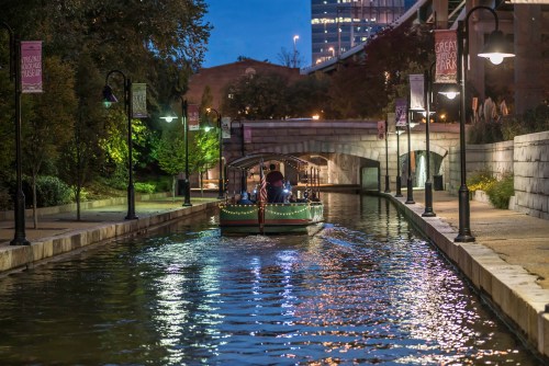 A boat glides through a softly lit canal, surrounded by trees and buildings, under a twilight sky.