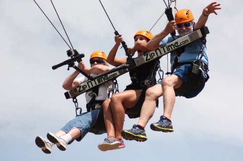 Three people in harnesses ziplining through the air, wearing helmets and smiling against a cloudy sky.