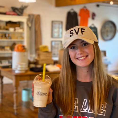 A smiling woman in a cap holds a smoothie in a cozy shop filled with various items.