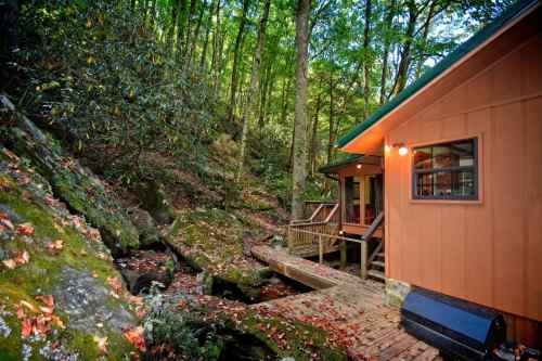 A cozy cabin nestled in a forest, surrounded by trees and autumn leaves, with a wooden deck and rocky landscape.
