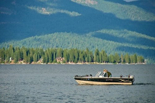 A boat glides across a calm lake, surrounded by lush green mountains and trees in the background.