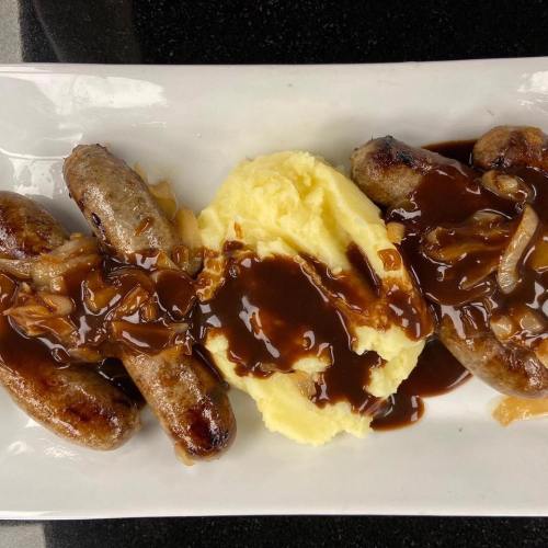 Three sausages topped with brown gravy and onions, served alongside a mound of creamy mashed potatoes on a white plate.