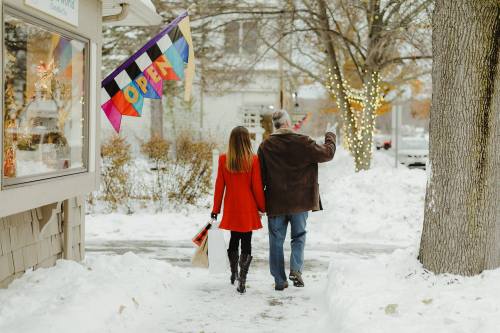 A couple walks hand in hand down a snowy street, passing an "OPEN" sign and festive decorations.
