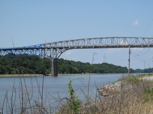 A blue and silver bridge spans a river, surrounded by greenery and a clear blue sky.