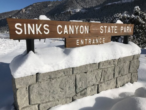 Wooden sign for Sinks Canyon State Park entrance, partially covered in snow, with mountains in the background.