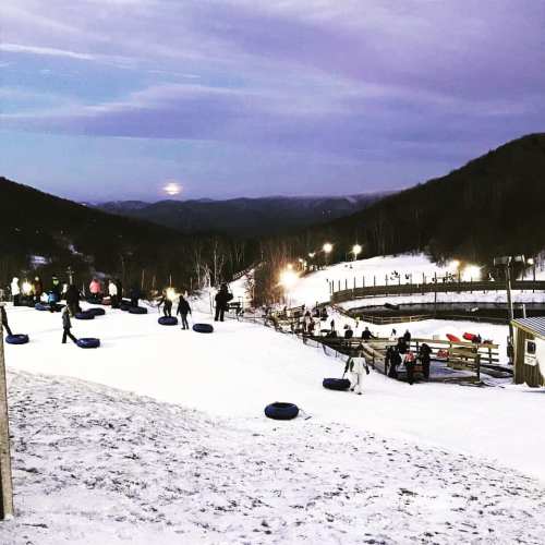 A snowy hill with people tubing, surrounded by mountains and a twilight sky. Lights twinkle in the background.