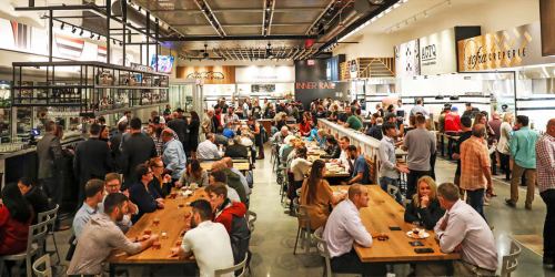 A bustling food hall filled with people dining at tables and enjoying various food options in a lively atmosphere.