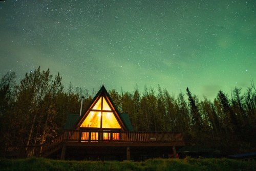 A cozy A-frame cabin illuminated at night, surrounded by trees under a starry sky with a hint of northern lights.