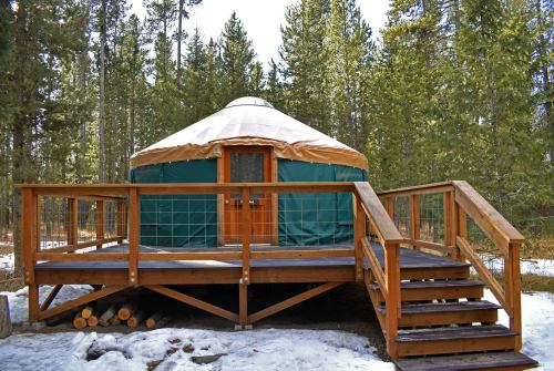 A green and tan yurt on a wooden deck, surrounded by trees and snow, with a staircase leading up to the entrance.