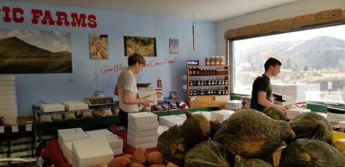 Two workers pack boxes in a farm store filled with produce and jars, with scenic mountains visible through the window.