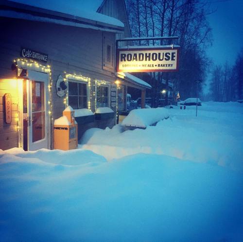 A cozy roadhouse with a sign, surrounded by deep snow and twinkling lights, in a wintery landscape.