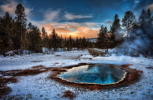 A steaming hot spring surrounded by snow and pine trees under a colorful sky at sunset.