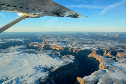 Aerial view of a snow-covered canyon with a plane wing in the foreground, under a clear blue sky.