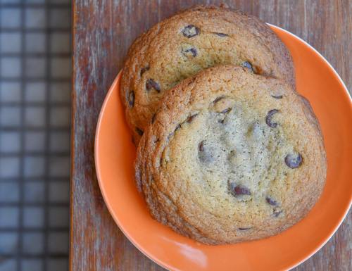 Two chocolate chip cookies on an orange plate, resting on a wooden surface.