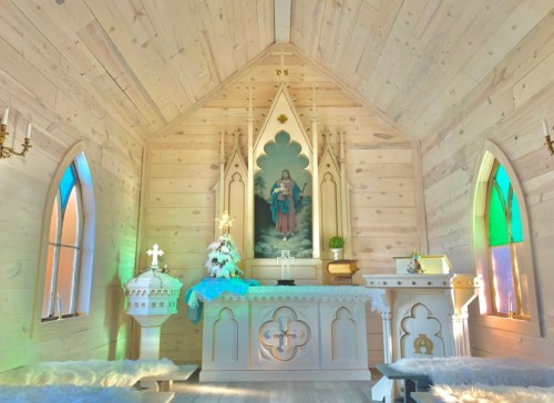 Interior of a wooden chapel featuring an altar, stained glass windows, and a Christmas tree. Soft lighting enhances the ambiance.