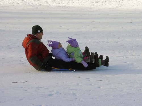 A man and two children sledding down a snowy hill, all wearing winter coats and hats, enjoying a fun day outdoors.