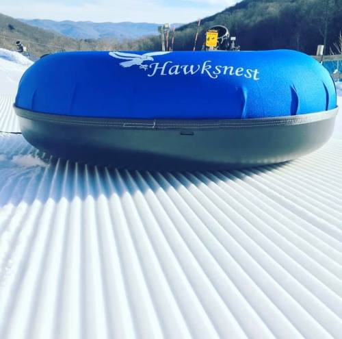 A blue snow tube with "Hawksnest" logo resting on a snowy slope, surrounded by mountains in the background.