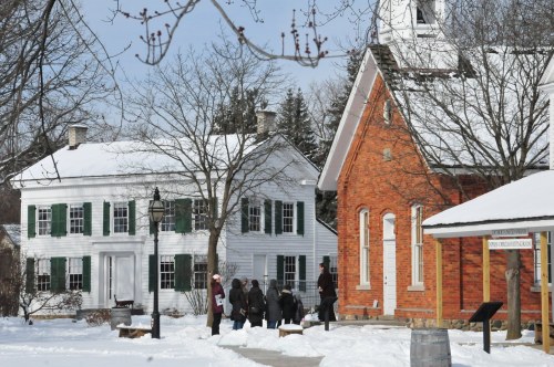 A snowy scene featuring a white house with green shutters and a red brick church, with people gathered outside.