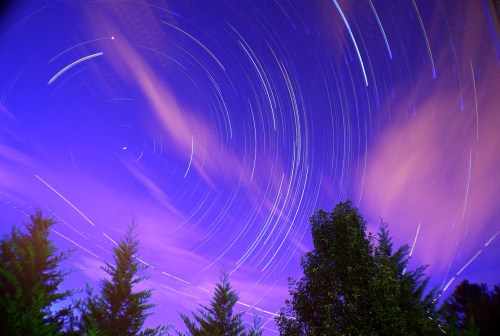 A starry night sky with swirling star trails and wispy clouds above silhouetted trees.