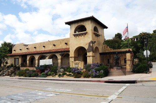 Historic building with a stucco facade, arched windows, and a flag, surrounded by greenery and flowers.