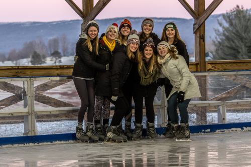 A group of eight women in winter attire smiles together on an ice skating rink, surrounded by a scenic winter landscape.