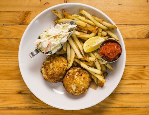 A plate with two crab cakes, French fries, coleslaw, a lemon wedge, and marinara sauce on a wooden table.