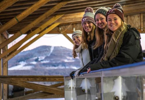 Four young women smile while leaning on a railing, with a snowy mountain landscape in the background.